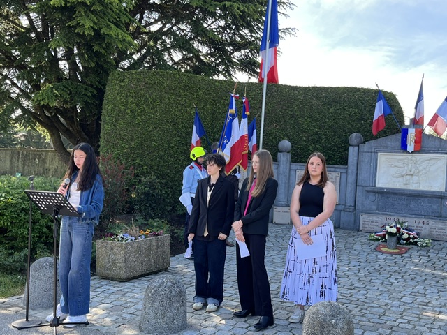 The image shows a group of people standing in front of a memorial, each holding a flag, under a clear sky with some trees and a cobblestone path.Le contenu généré par l’IA peut être incorrect.